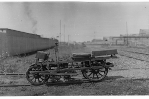 Ein altes Schwarz-Weiß-Foto eines Motorrads auf der Bahn, umgeben von Gras, Pflanzen, einer Wand, Pfählen, Gebäuden und einem Himmel im Hintergrund.