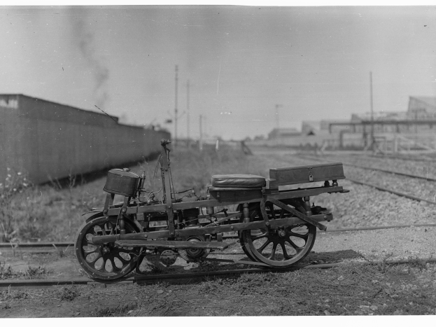 Ein altes Schwarz-Weiß-Foto eines Motorrads auf der Bahn, umgeben von Gras, Pflanzen, einer Wand, Pfählen, Gebäuden und einem Himmel im Hintergrund.