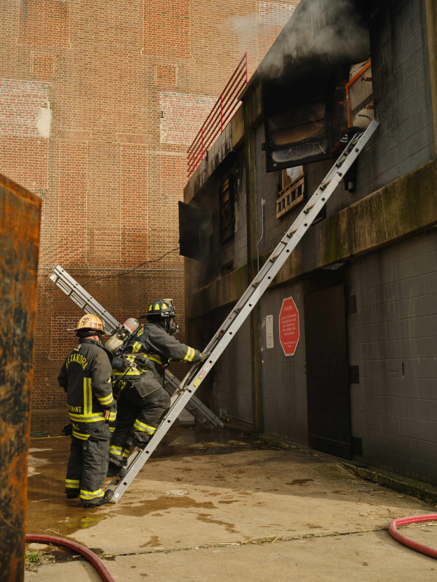 Feuerwehrleute in Helmen klettern an einer Leiter zu einem rauchgefüllten Gebäude hoch, mit einem Metallobjekt links und sichtbaren Rohren unten.