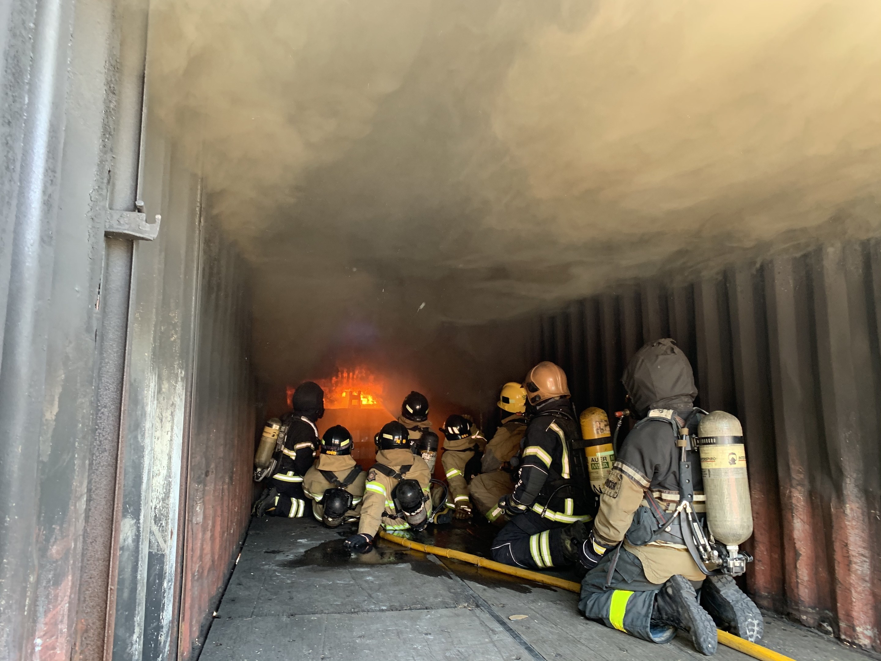 Feuerwehrleute in Schutzanzügen und Sauerstoffflaschen arbeiten in einem raucherfüllten Tunnel mit einem sichtbaren Feuer im Hintergrund, einer hält einen Schlauch.