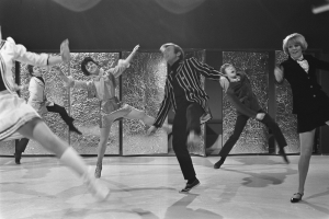Eine Gruppe von Menschen, die auf einer Bühne tanzen, mit einer Wand im Hintergrund, alle in Schwarz-Weiß und mit Schuhen.