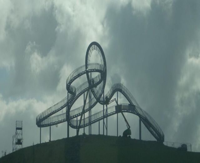 Eine Achterbahn auf dem Gipfel eines Hügels mit einem bewölkten Himmel, umgeben von Stützpfählen und -strukturen.