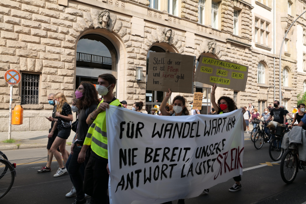 Eine Gruppe von Menschen marschiert bei einer Protestaktion gegen die Anti-Kriegsbewegung in Berlin, einige halten Schilder und andere fahren Fahrräder, im Hintergrund ein Gebäude mit Fenstern, Bogengängen, Säulen und Skulpturen.