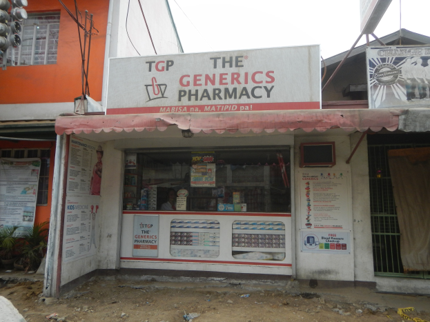 Ein Apotheken-Schild mit der Aufschrift "The Generics Pharmacy" vor einem Gebäude mit umliegenden städtischen Elementen unter einem klaren blauen Himmel.