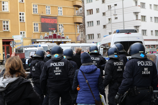 Polizeibeamte in Uniform stehen vor einer Menge mit Helmen und Jacken während einer Demonstration in Berlin, Deutschland, mit Fahrzeugen, Gebäuden, Laternenpfählen und einem Banner im Hintergrund.