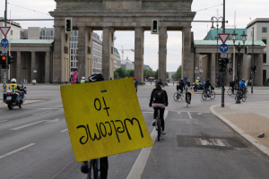 Eine Gruppe von Radfahrern in Helmen fährt eine Straße vor dem Brandenburger Tor in Berlin, Deutschland, entlang, einer hält ein gelbes Schild, unter einem klaren blauen Himmel mit Gebäuden und Bäumen im Hintergrund.