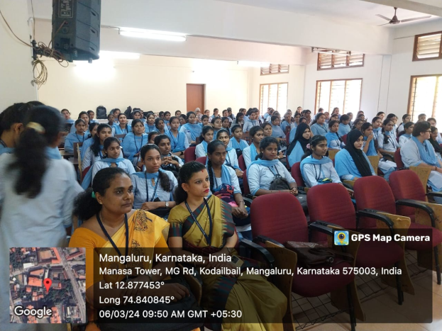 Gruppe von Menschen, die in einem Raum mit Fenstern, Lampen, einem Ventilator und einer Karte mit Text unten sitzen und einem Redner zuhören.
