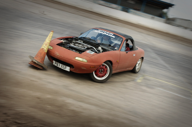 Eine Person in einem roten Auto auf einer Rennstrecke mit einem Verkehrskegel vorne, der Hintergrund durch hohe Geschwindigkeit unscharf.