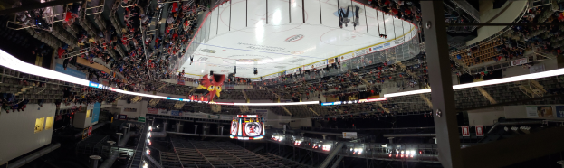 Eine große Arena voller Zuschauer bei einem Eishockeyspiel mit Stühlen, Geländern, Tafeln mit Text, Lampen, Pfosten und einer Treppe, die zu den Sitzplätzen führt.
