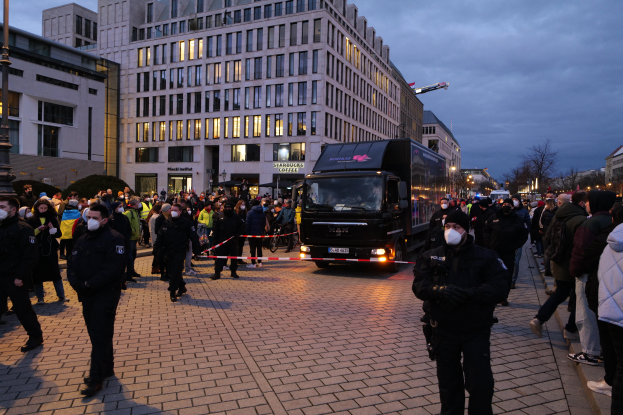 Eine Gruppe von Menschen steht vor einem Lastwagen auf einer Straße, umgeben von Gebäuden, Laternenpfählen, Bäumen und einem bewölkten Himmel, wobei einige Mützen und Masken tragen und ein Band mit einem Pfahl im Vordergrund zu sehen ist.