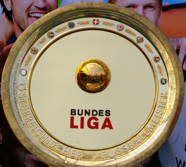 Ein Mann hält einen goldenen Schild mit der Aufschrift 'Bundesliga', im Hintergrund ein Banner mit drei Personen.