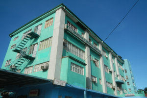 Ein großes grünes Gebäude mit vielen Fenstern, Klimaanlagen und Treppen, umgeben von Bäumen, Strommasten und Fahrzeugen auf der Straße unter einem klaren blauen Himmel.
