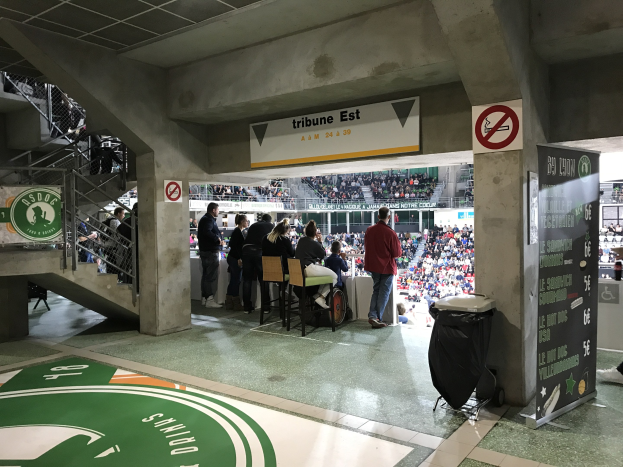 Eine Gruppe von Menschen steht vor einem sitzenden Publikum in einem Stadion, mit Schildern, die "Boston Celtics vs New York Jets" anzeigen, einem Müllcontainer, einer Tafel mit Text, einer Treppe mit Geländern und einer Decke mit Deckenleuchten.