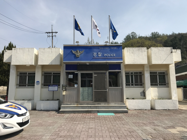 Eine Polizeistation mit einem geparkten Auto, Fenstern, einer Tür und einem Schild, das Flaggen, einen Strommast und Bäume im Hintergrund unter einem bewölkten Himmel zeigt.