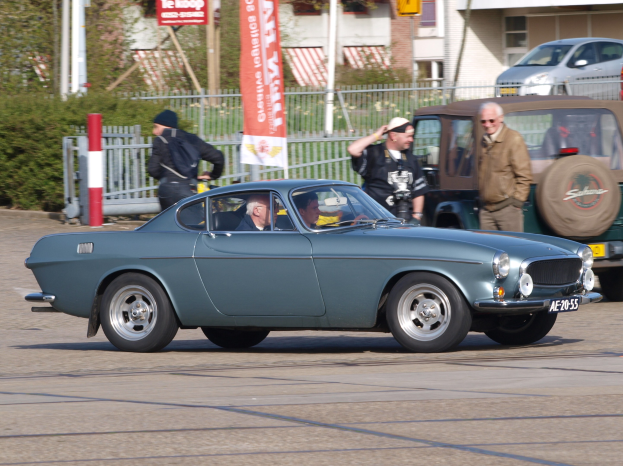 Eine Gruppe von Autos fährt auf einer Straße mit Passagieren, flankiert von Gebäuden, Bäumen und verschiedenen Straßenobjekten, über einer sichtbaren Straße.