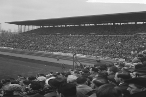 Schwarzes und weißes Foto eines vollen Stadions mit Zuschauern, die ein Fußballspiel verfolgen, einige stehen und andere sitzen auf Tribünen, mit Schildern, Pfählen, einer Hütte, Bäumen, einem Turm und einem bewölkten Himmel im Hintergrund.