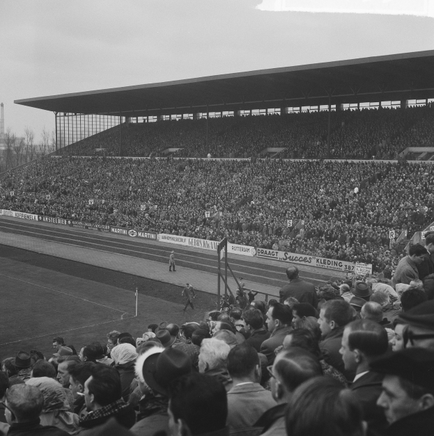Schwarzes und weißes Foto eines vollen Stadions mit Zuschauern, die ein Fußballspiel verfolgen, einige stehen und andere sitzen auf Tribünen, mit Schildern, Pfählen, einer Hütte, Bäumen, einem Turm und einem bewölkten Himmel im Hintergrund.