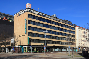 Ein großes Gebäude mit zahlreichen Fenstern, identifiziert als das Fore Inn Hotel Berlin, umgeben von Straßeninfrastruktur einschließlich Pfosten, Lampen, Signalen, Schildern, Bäumen und einem bewölkten Himmel.