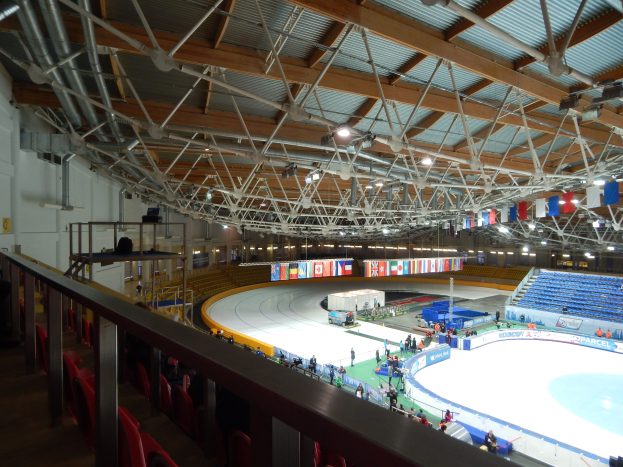 Großes Innen-Eisstadion mit zahlreichen Eisläufern, umgeben von Sitzplätzen, Geländern, Brettern, Beleuchtung und konstruktiven Elementen, mit einer Wand im Hintergrund.