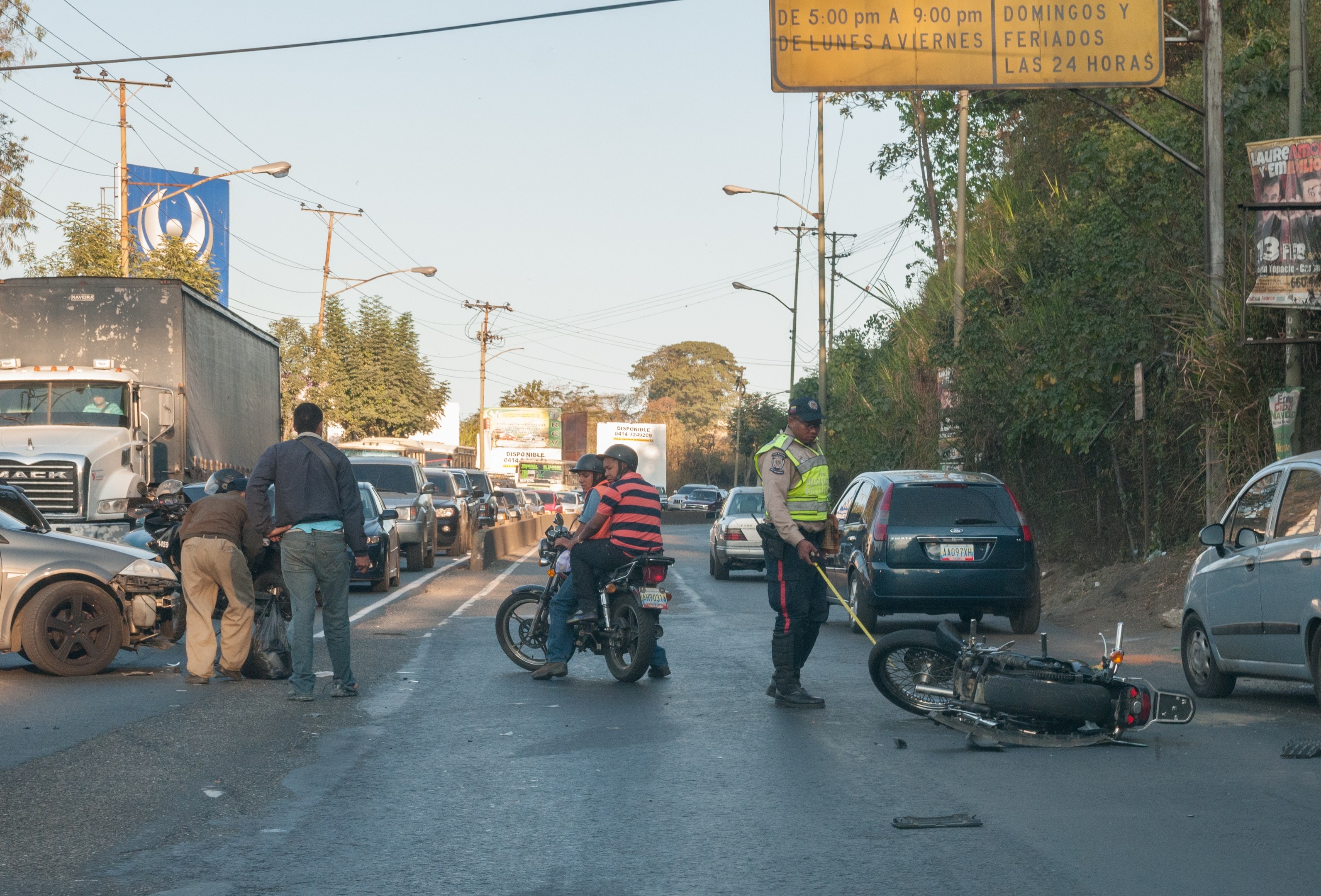 Gruppe von Menschen um ein verunglücktes Motorrad auf der Straße mit mehreren Fahrzeugen, darunter ein Lastwagen, und Hintergrundelementen wie Bäumen, Pfählen, Lampen, Schildern und Himmel.