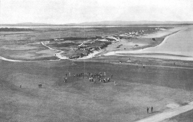 Schwarz-weiß-Foto von Golfern auf dem 18. Loch am Royal Birkham Golf Club, mit saftig grünem Rasen, verstreuten Häusern, sanften Hügeln und einem hellblauen Himmel im Hintergrund.