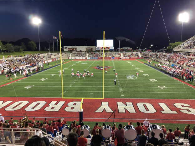 Ein nächtliches Fußballspiel in einem Stadion mit Spielern auf dem Feld, Zuschauern auf Stühlen und einer Band, die Instrumente spielt, umgeben von einem Metallzaun und Hintergrundelementen wie Laternen, Fahnen, einem Anzeigebildschirm, Bäumen, Hügeln und einem sternenklaren Himmel.
