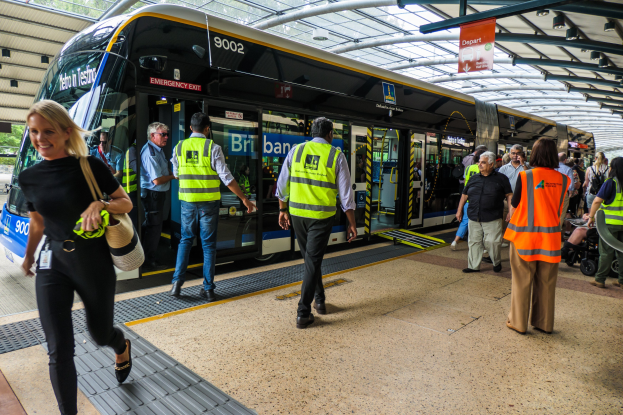 Eine Gruppe von Menschen geht auf einen Zug an einem Bahnhof zu, mit einer Frau im Vordergrund, die eine Handtasche trägt, und einem Mann im Hintergrund, unter einem Dach, das von Metallpfosten gestützt wird und ein Banner mit Text trägt.