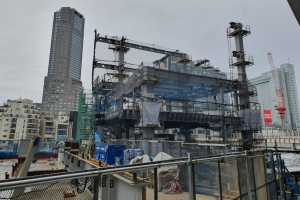 Großer Bauplatz für ein neues Kraftwerk in einer städtischen Gegend, umgeben von einem Metallzaun, mit sichtbaren Gebäuden, Fahrzeugen, Pfählen, Drähten, einem Banner und einem bewölkten Himmel.