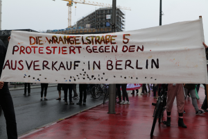 Eine Gruppe von Menschen steht auf einer Straße und hölt eine Plakate mit der Aufschrift "Die Wrangellstraße 5 Protestiert Gegen den Ausverkauf in Berlin", mit Fahrrádern, Masten, Gebäuden und einem Kran im Hintergrund und einem sichtbaren Himmel.