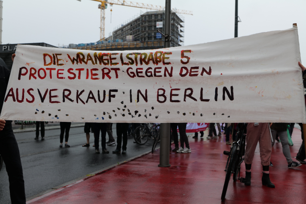 Eine Gruppe von Menschen steht auf einer Straße und hölt eine Plakate mit der Aufschrift "Die Wrangellstraße 5 Protestiert Gegen den Ausverkauf in Berlin", mit Fahrrádern, Masten, Gebäuden und einem Kran im Hintergrund und einem sichtbaren Himmel.