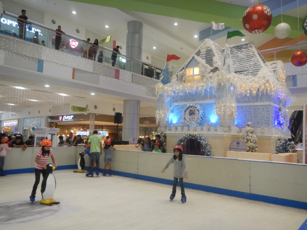 Menschen, die auf einer Eisbahn in einem Einkaufszentrum Schlittschuh laufen, umgeben von festlichen Däcorations wie einem häusähnlichen Gebäude mit Lichtern und einer Menge, die von Geländern und Säulen beobachtet.