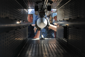 Zwei Männer in Helmen und Handschuhen arbeiten an einer Maschine in einem Raum, halten Gegenstände und sind von Metallkomponenten umgeben.