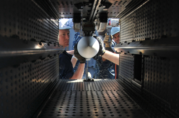 Zwei Männer in Helmen und Handschuhen arbeiten an einer Maschine in einem Raum, halten Gegenstände und sind von Metallkomponenten umgeben.