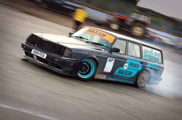 Ein Auto auf einer Rennstrecke, das Rauch ausstößt, umgeben von anderen Fahrzeugen und Zuschauern, mit einem unscharfen Hintergrund, der hohe Geschwindigkeit andeutet.