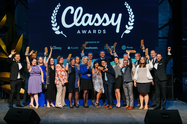 Eine Gruppe von Menschen auf einer Bühne, die strahlen und Preise halten, mit Lautsprechern unten und einem Bildschirm im Hintergrund, der "Classy Awards 2019" anzeigt.