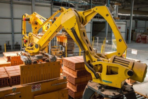 Großes industrielles Lagerhaus mit gestapelten Kisten, Kränen an der Decke, Zaun, Versorgungsmasten und einer fernen Wand in einer automatisierten Fertigungsumgebung.