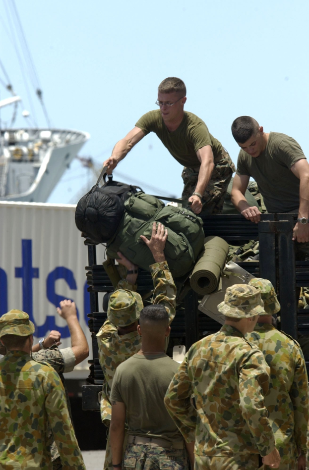 Männer in Militäruniformen laden Gepäck auf einen Lastwagen mit einem Schiff und einem Container im Hintergrund unter einem klaren blauen Himmel.