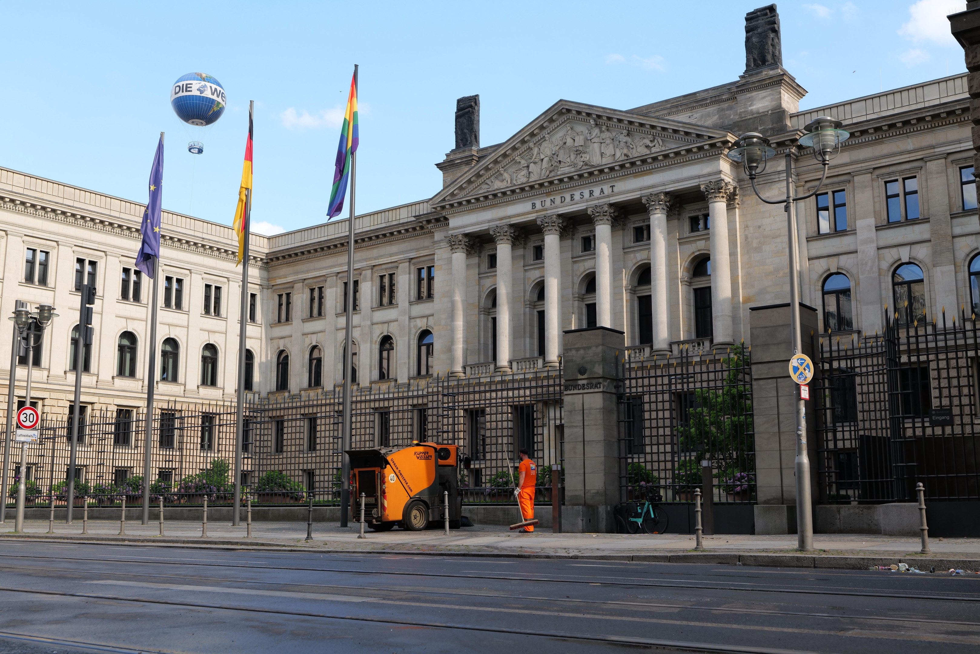 Großes modernes Gebäude mit zahlreichen Fenstern, als Bundestag in Berlin, Deutschland identifiziert, umgeben von Straßeninfrastruktur, Fußgängern, Fahrzeugen und Flaggen, unter einem bewölkten Himmel.