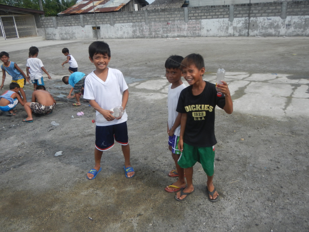Junge Jungs in einer Parklücke mit Flaschen in der Hand, mit Häusern und Bäumen im Hintergrund unter einem klaren blauen Himmel.