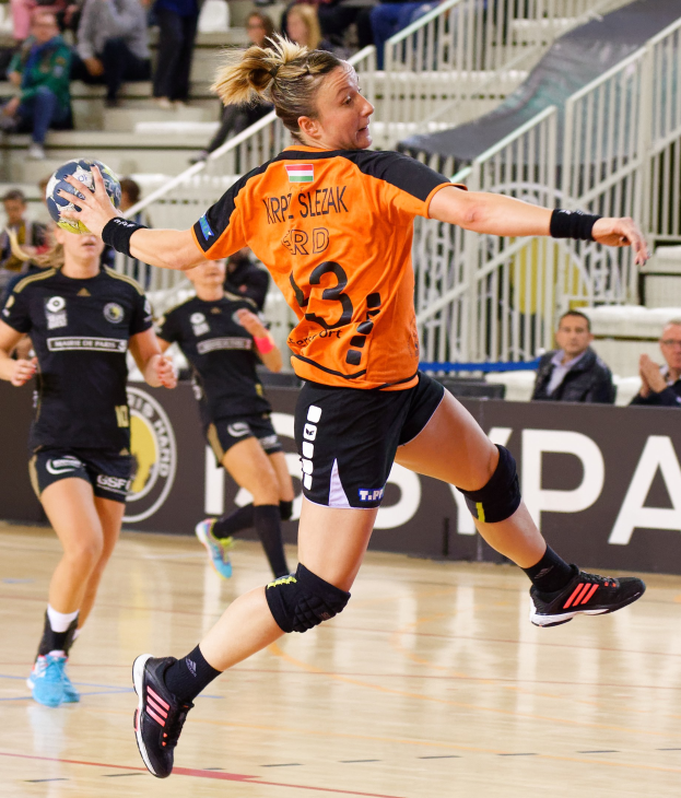 Eine Frau in einem orangen T-Shirt und schwarzen Shorts hält einen Handball, während Zuschauer auf Treppen und Geländern hinter ihr sitzen.