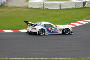 Ein Porsche 911 GT3 RSR Rennauto auf einer Rennstrecke, umgeben von grünem Gras und einem Zaun auf der linken Seite, mit Text und Zahlen, die die Teilnahme an den 24 Stunden von Le Mans anzeigen.