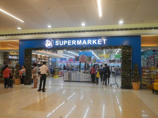 Eine Gruppe von Menschen, die vor einem Supermarkt stehen, einige halten Einkaufstüten, mit einem Einkaufswagen, einem Topf mit Pflanze, Regalen mit Gegenständen, Schildern und einer Decke mit Leuchten.