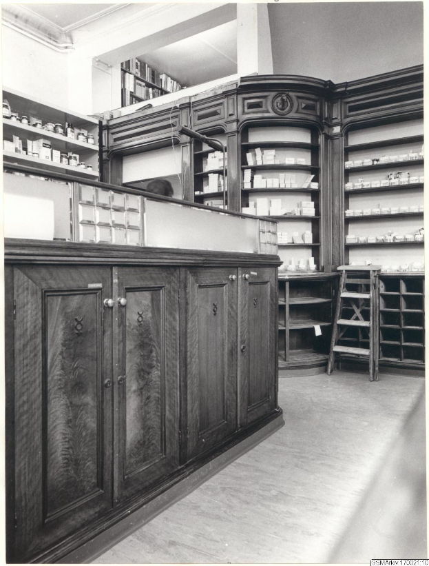 Schwarz-weiß-Foto eines vintage-Apotheken-Interieurs mit einer Theke, Schränken, einer Leiter, Regalen und Text oben.