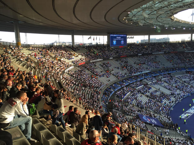 Große Menschenmenge in einem Stadion bei einem Fötballspiel, mit einer Bühne auf der rechten Seite, Fahnen, Masten, einem Bildschirm und der Allianz Arena in München, Deutschland im Hintergrund.