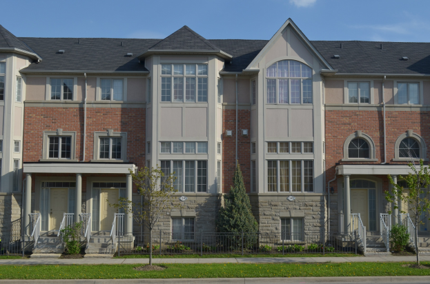 Eine Reihe von Stadthäusern in einer Wohngegend mit Fenstern, Türen, Säulen, Geländern, Stufen, Pflanzen, Bäumen, Gras, einer Straße im Vordergrund und dem Himmel im Hintergrund.