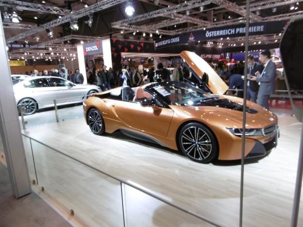 Ein elegantes schwarzes BMW i8 Cabrio mit Chromakzenten, das eine lange Motorhaube, einen tiefgelegten Körper, eine breite Kühlergrill und Scheinwerfer aufweist, das auf der IAA in Frankfurt umgeben von formell gekleideten Gästen in einer Atmosphäre der Aufregung gezeigt wird.