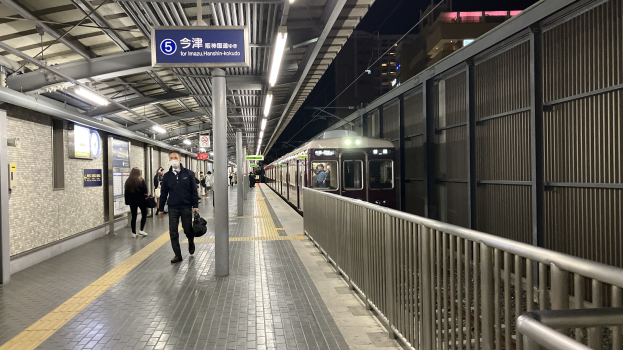 Ein Zug fährt auf einem Bahnsteig ein, mit Menschen in der Nähe, einem Geländer auf der rechten Seite, Gebäuden im Hintergrund, Bahnsteiglampen und einer Abfahrtsanzeige.