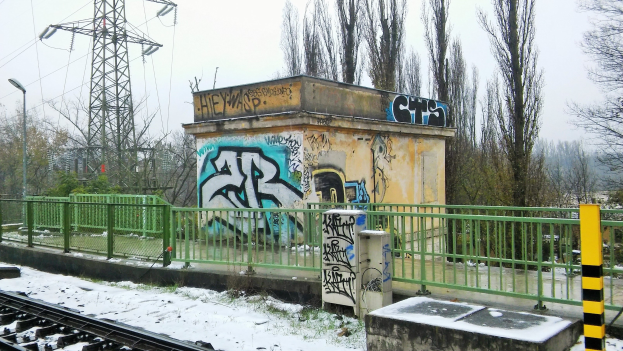 Eisenbahnschiene mit Graffiti an der Seite, umgeben von Geländern, Pfählen, Lampen und Bäumen, mit Türmen und Drähten im Hintergrund und einem klaren Himmel und Schnee am Boden.