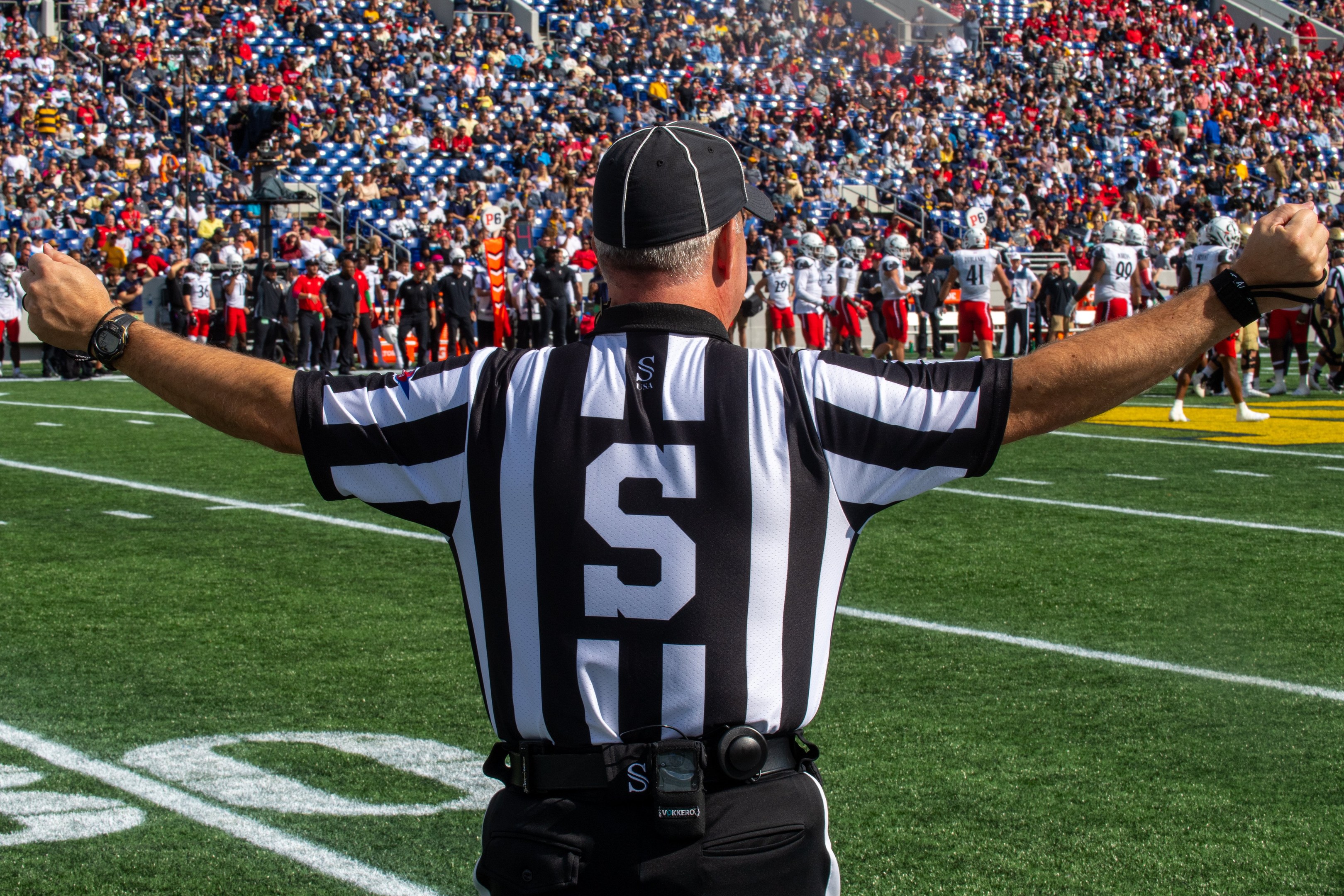 Ein Schiedsrichter steht auf einem Fußballfeld mit ausgestreckten Armen, umgeben von Menschen auf dem Boden und einer sitzenden Menge im Hintergrund.