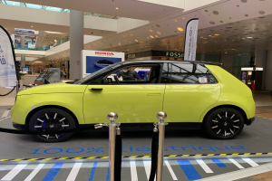 Ein gelbes wasserstoffbetriebenes Elektroauto vor einem Gebäude geparkt, umgeben von Absperrpollern mit Seilen, Bannern und Schildern, mit einer Person im Auto, einer Gruppe von Menschen in der Nähe, einer Säule, einem Topfpflanze, Deckenleuchten und einer Decke mit Deckenventilatoren.